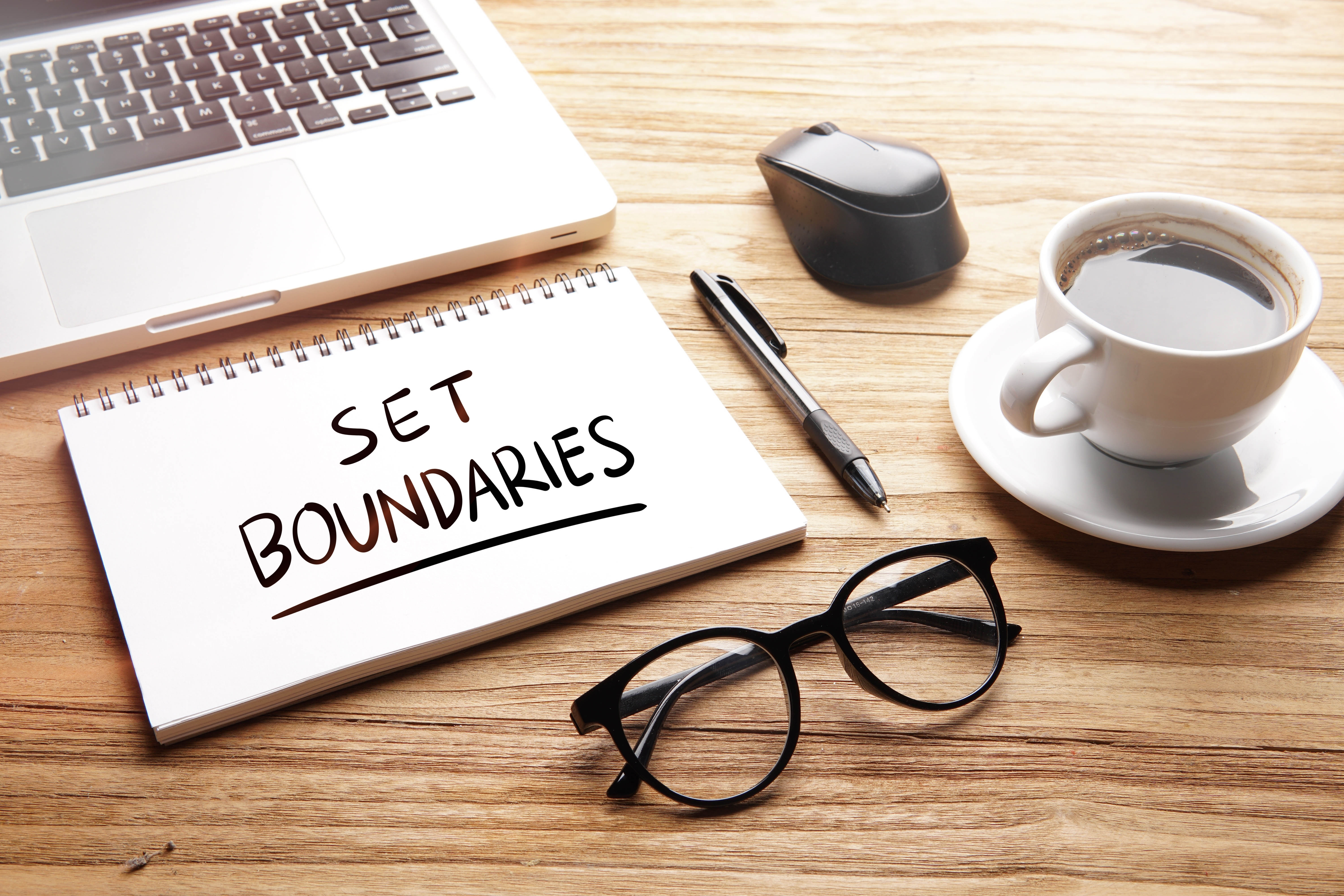 Close-up of a desk with a laptop and a notebook that says "Set boundaries" with glasses and coffee cup Close-up of a desk with a laptop and a notebook that says "Set boundaries" with glasses and coffee cup